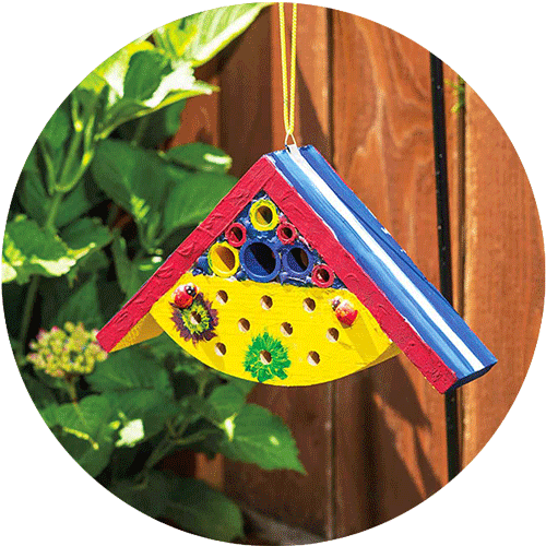 Red, yellow, green and blue painted hanging bug house with a shrub and fence in the background.